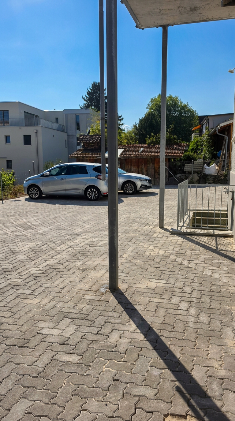 Frisch verlegter Verbundsteinplatz unter Betondach mit Metallpfosten und geparkten Autos